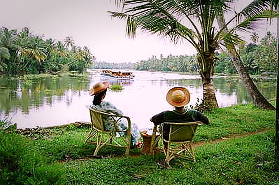 Two guests in garden chairs by the water, watching a houseboat go by